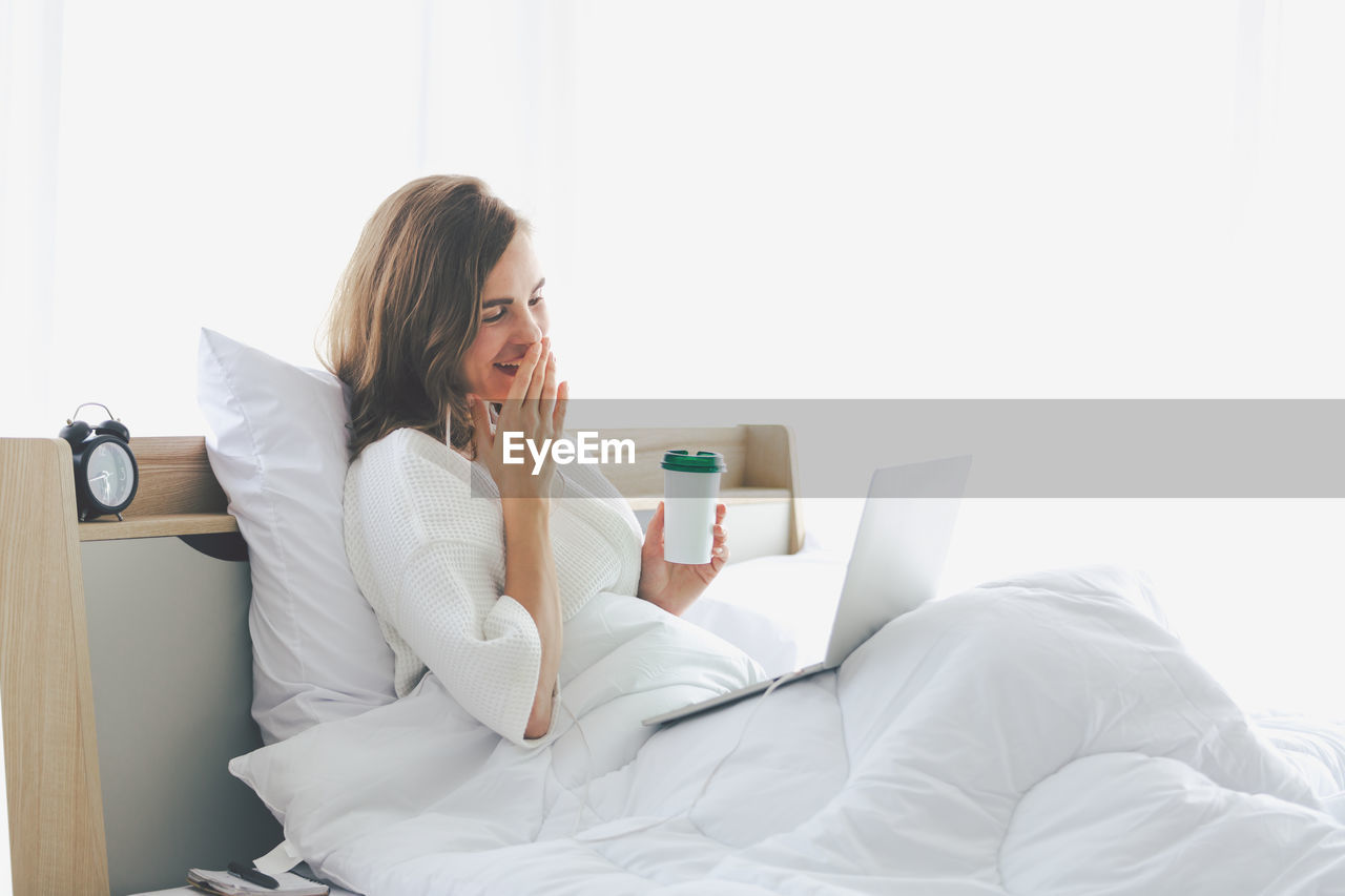 Young woman using laptop while holding coffee cup on bed at home