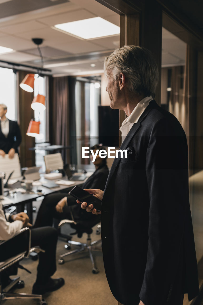 Senior male professional standing on doorway looking at colleagues during meeting board room