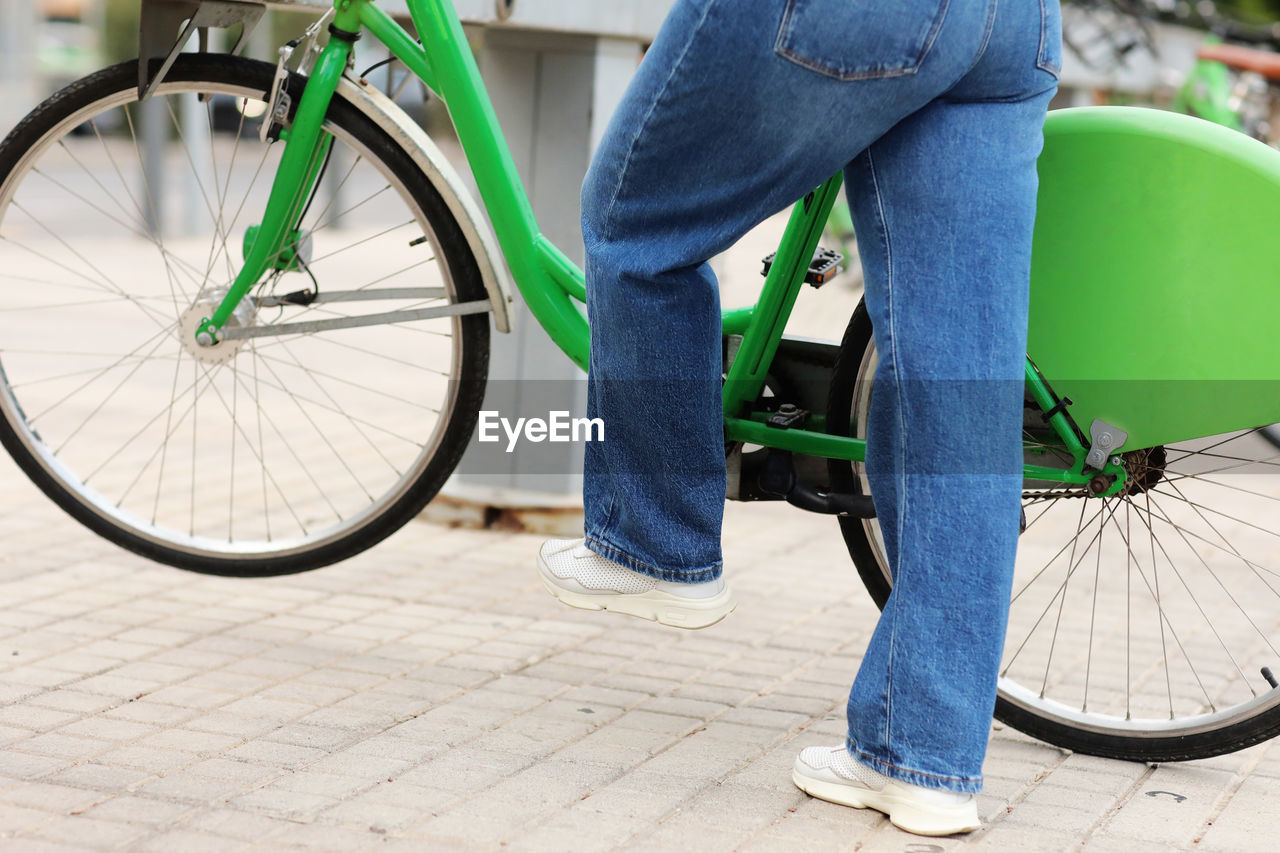 Cropped photo of unrecognizable woman in blue jeans is picking up a green bike at a bike rental 