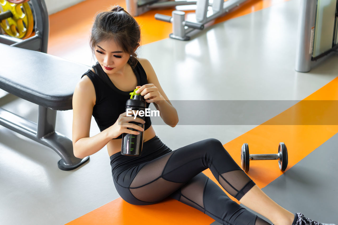 Young woman holding water bottle while sitting in gym