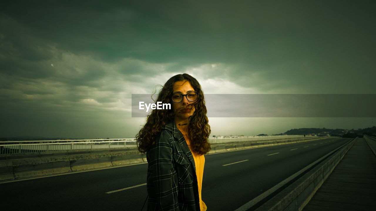 Portrait of young woman standing on road against cloudy sky