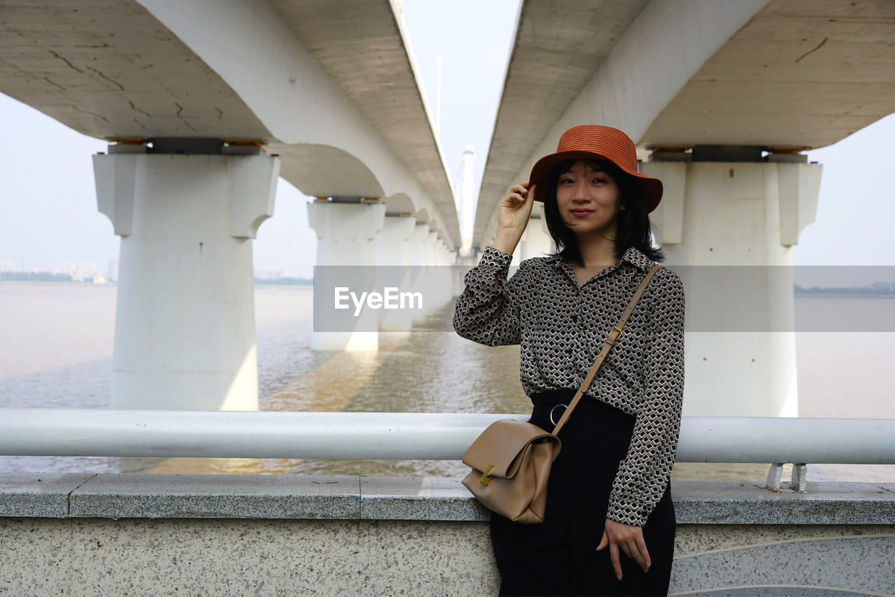 Portrait of beautiful young woman standing against bridge