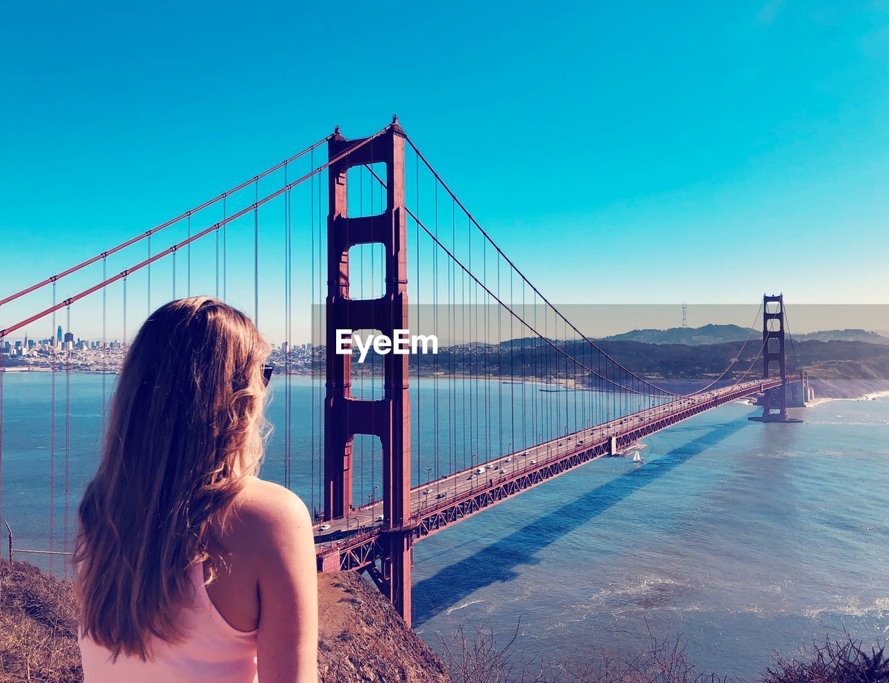 Rear view of young woman looking golden gate bridge over bay against clear blue sky