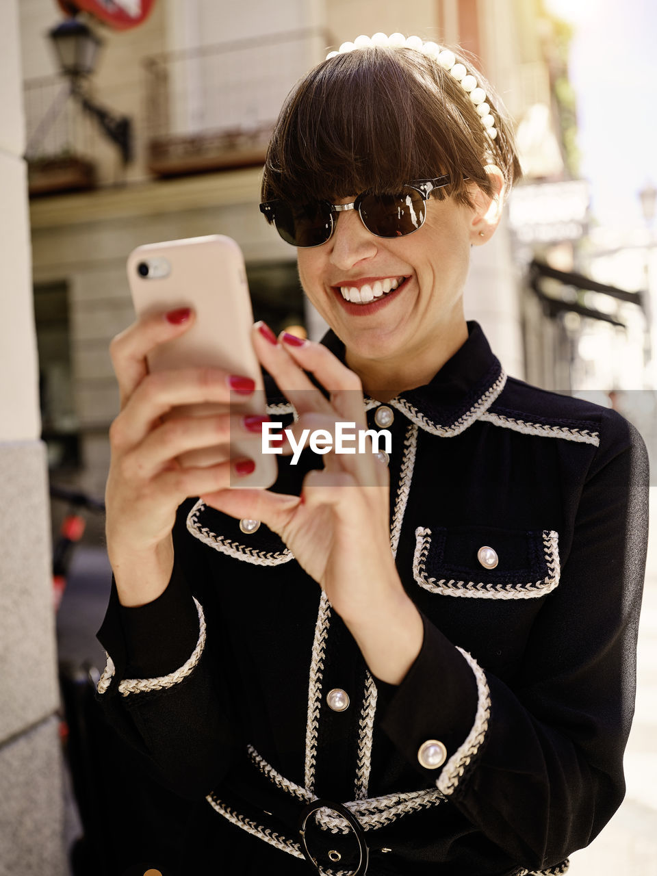 Smiling female with bob hairstyle and in trendy jacket and blouse standing on sidewalk on sunny day and messaging online via cellphone