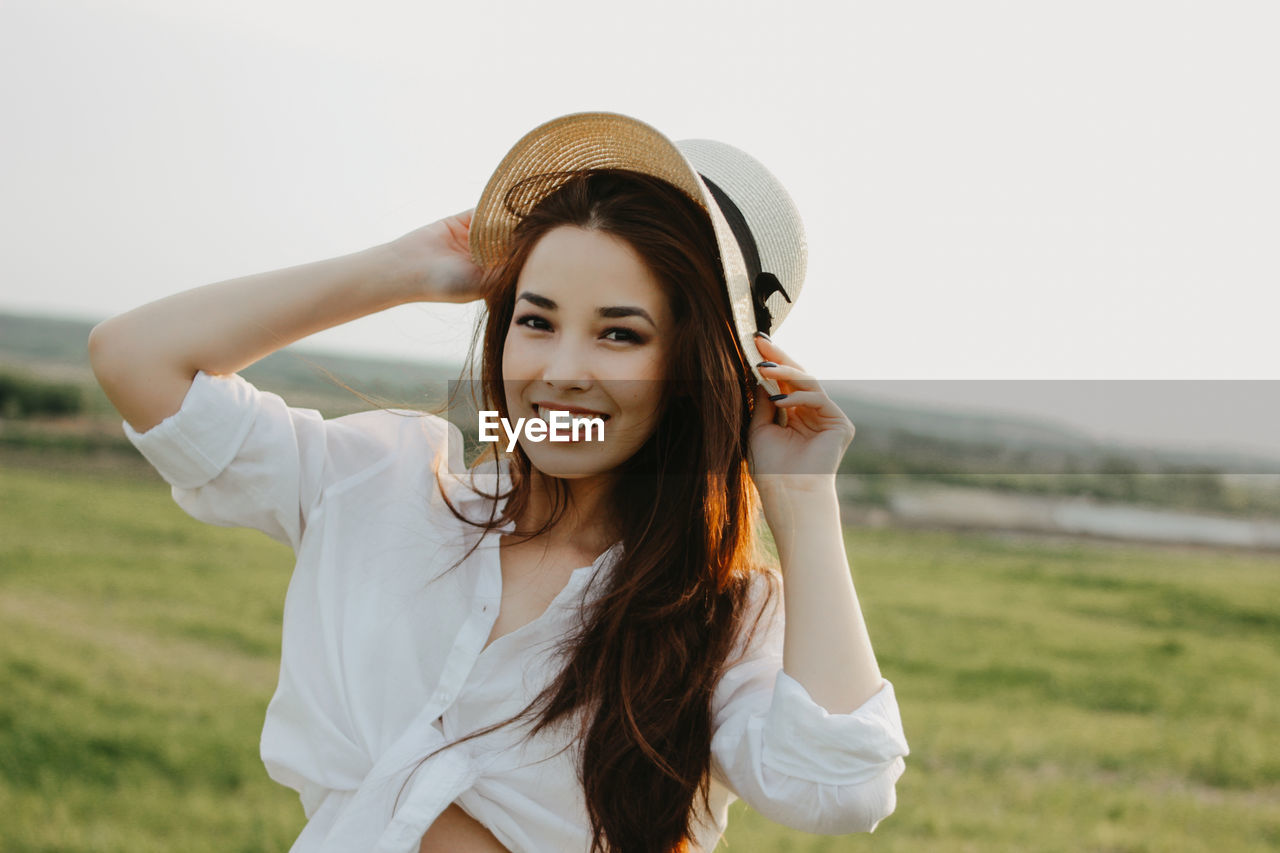 Portrait of smiling young woman with hat standing on land against clear sky