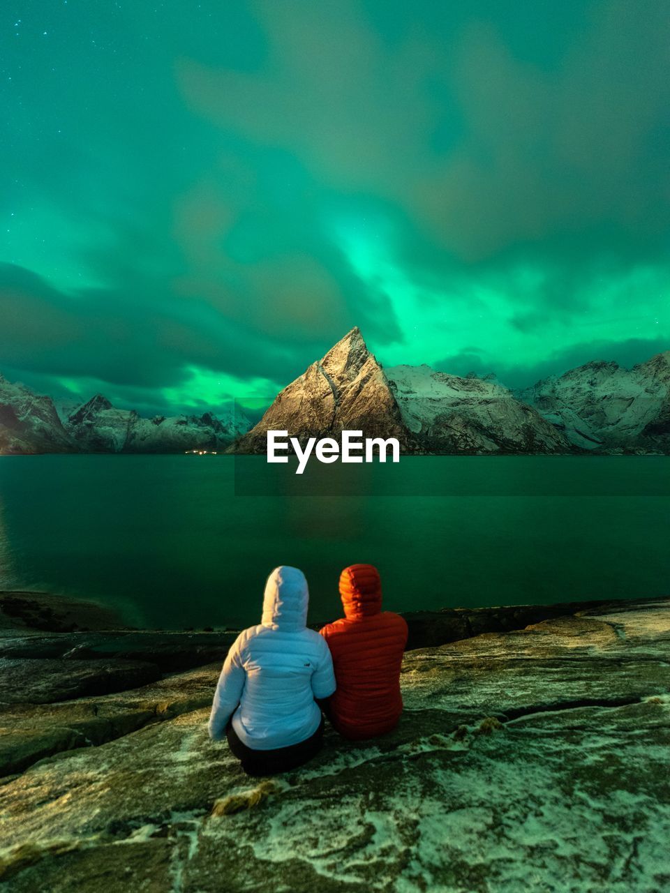 Rear view of hikers sitting on rock by lake against aurora