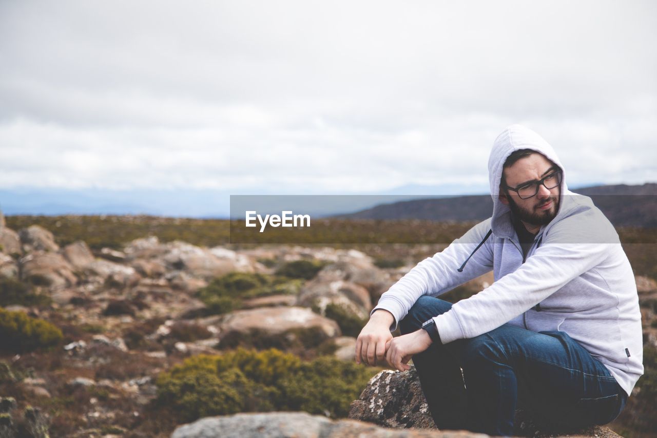 Young man looking away while sitting on rock against sky