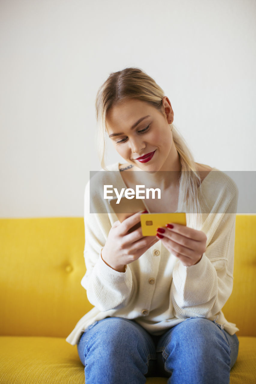 Blonde woman using smartphone and using bank card at home