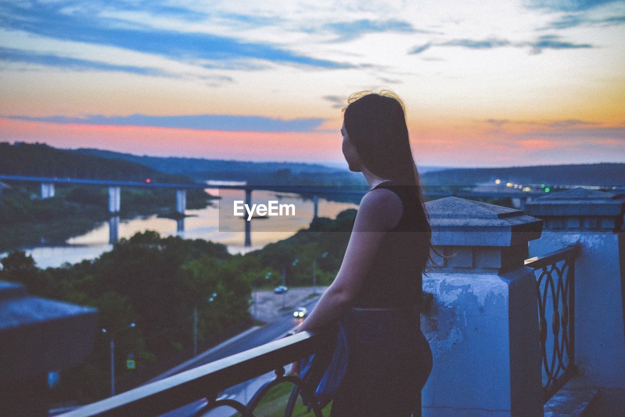 Young woman standing by railing against sky during sunset