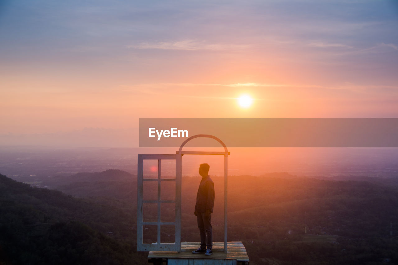 MAN STANDING ON MOUNTAIN AGAINST SKY AT SUNSET