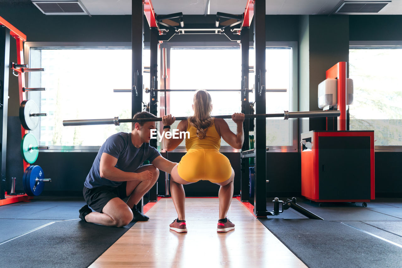 Back view of unrecognizable female athlete doing squats with heavy barbell during training in gym under supervision of male personal trainer