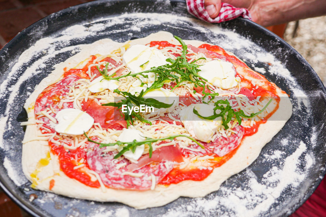 Raw pizza on baking tray with arugula prosciutto and cheese. 