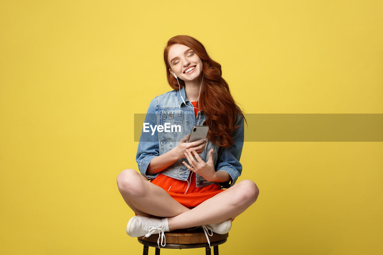 Woman listening music while sitting on stool against yellow background