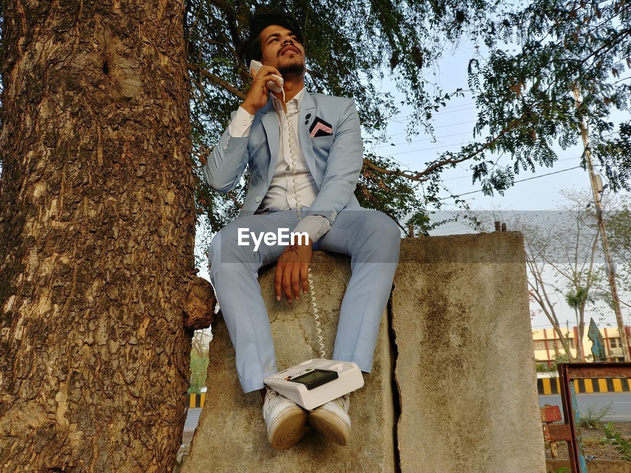 YOUNG MAN SITTING ON TREE TRUNK AGAINST PLANTS