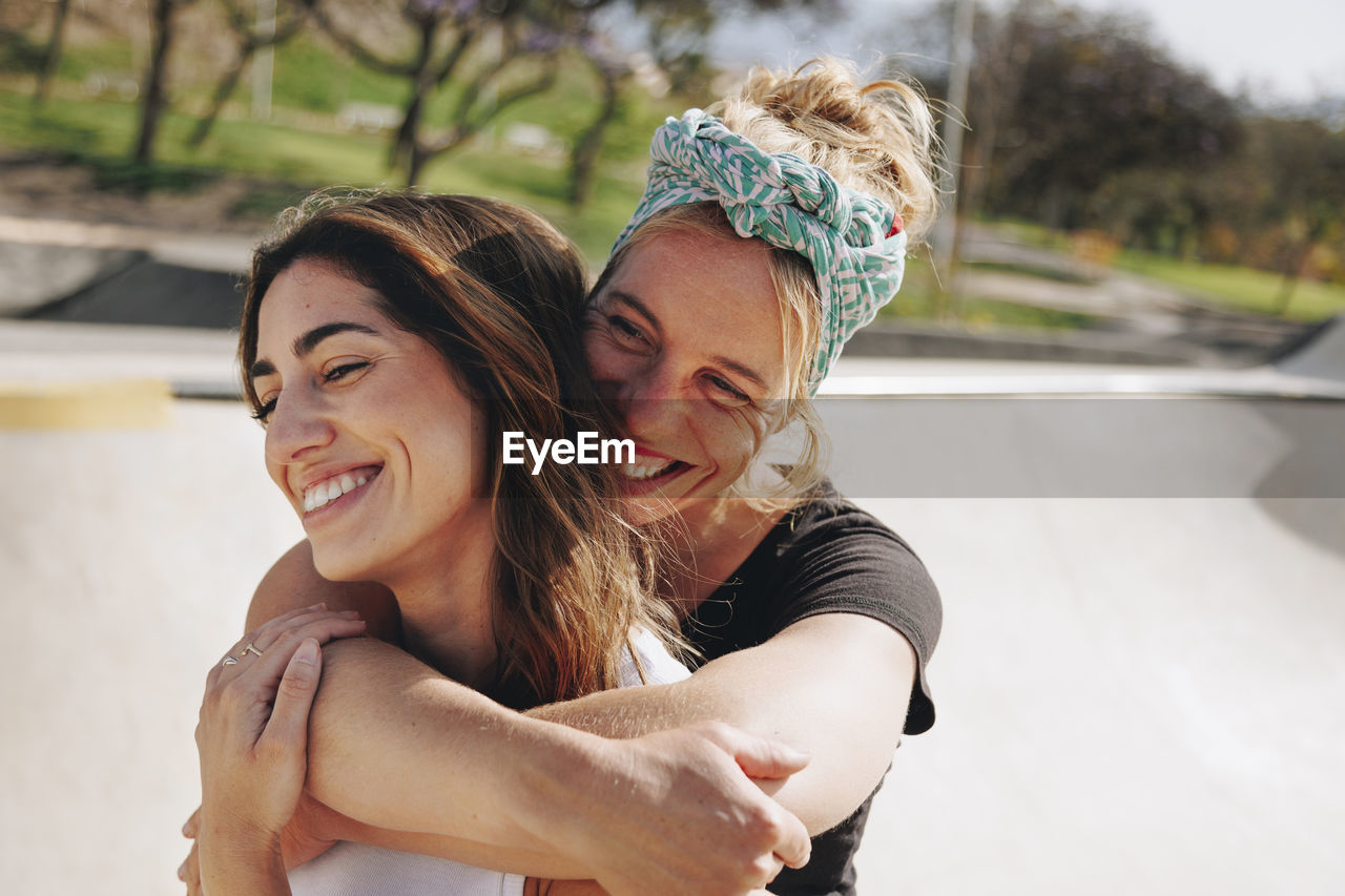 Smiling woman embracing friend from behind on sunny day