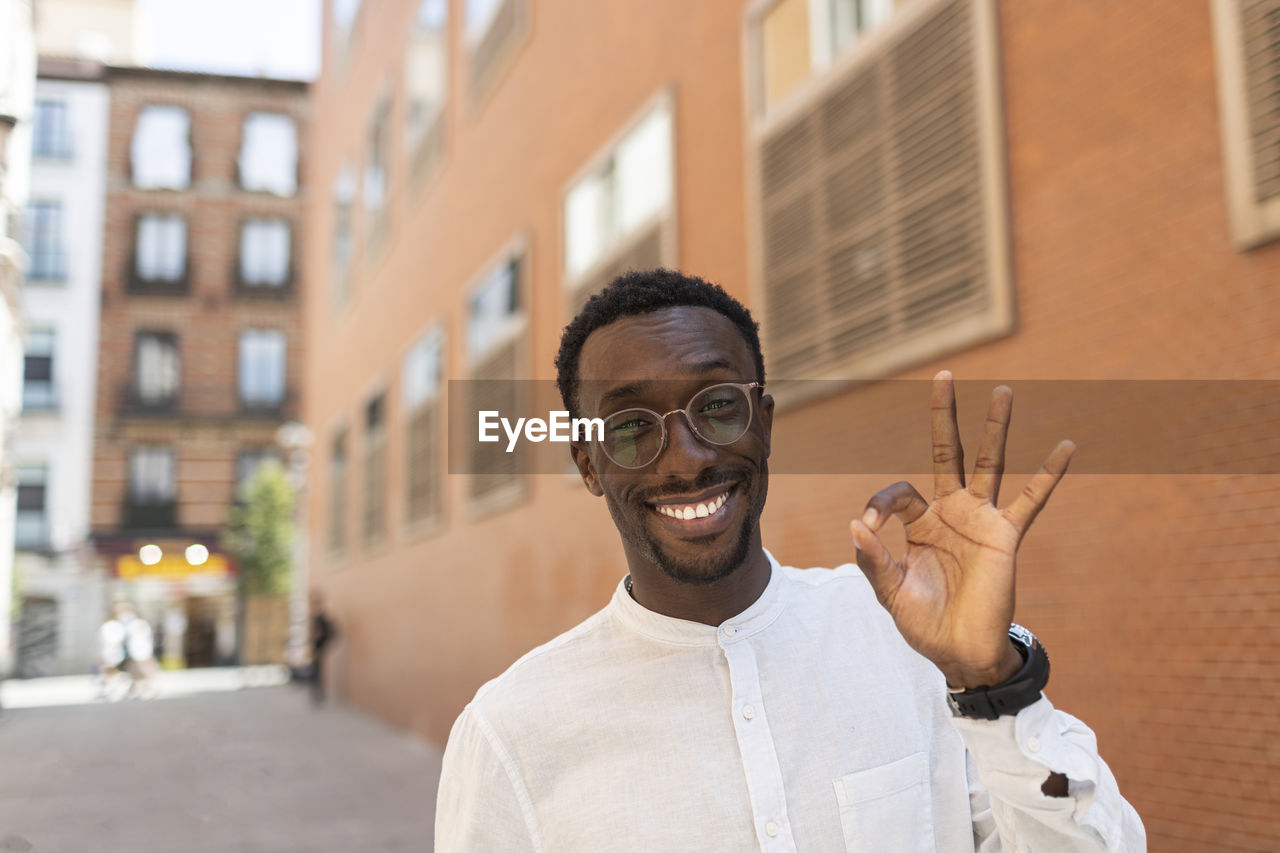 Smiling young man gesturing ok sign in front of buildings