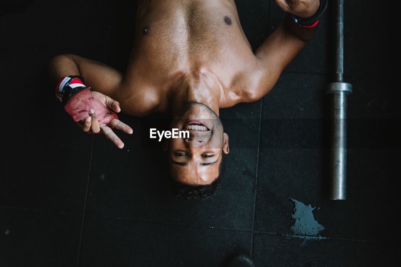 High angle portrait of shirtless man gesturing peace sign while lying on floor