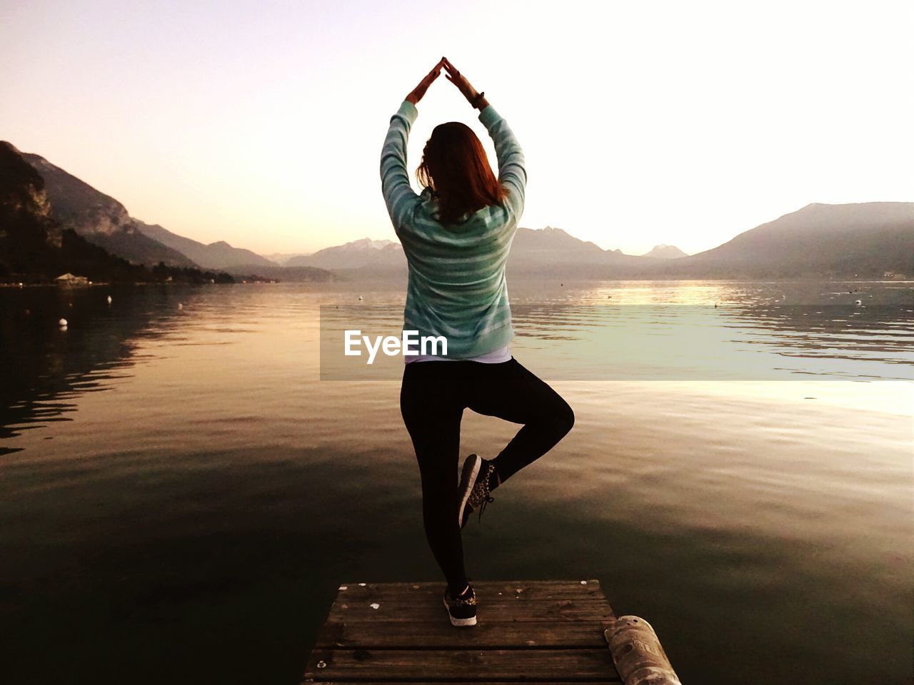 Rear view of woman doing yoga on pier over lake against sky during sunrise