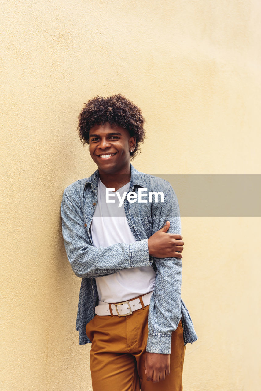 Determined african american male in trendy outfit and with afro hairstyle leaning on stone wall of building and looking at camera
