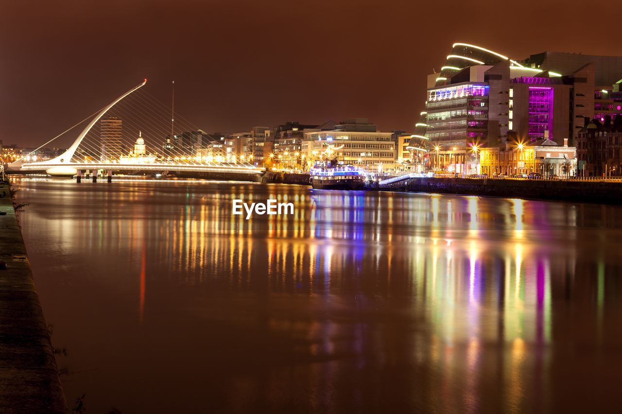 ILLUMINATED BRIDGE OVER RIVER BY BUILDINGS AGAINST SKY