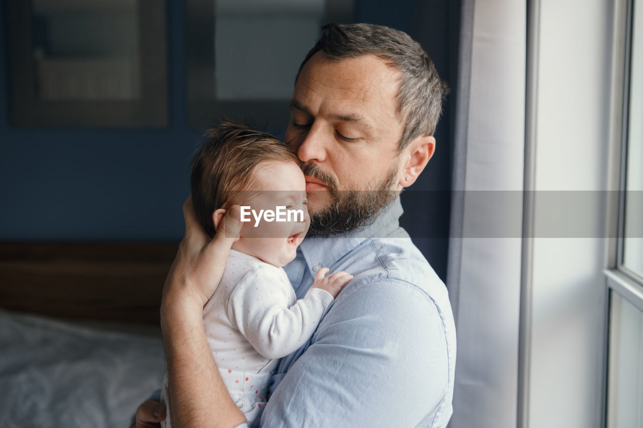 Side view of man playing with daughter while holding by window