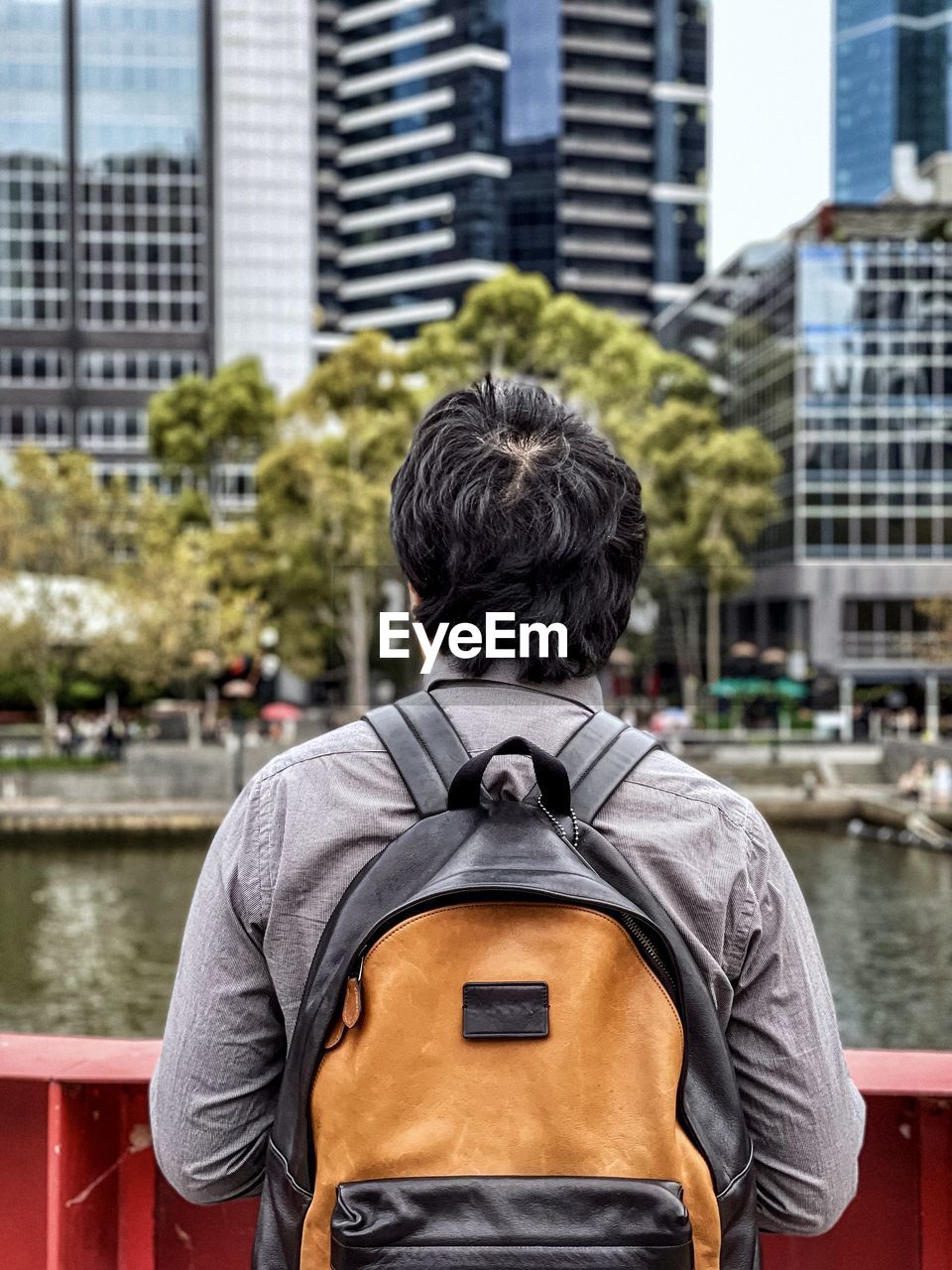 Rear view of man standing at metal fence and looking at river and skyscrapers in the city.