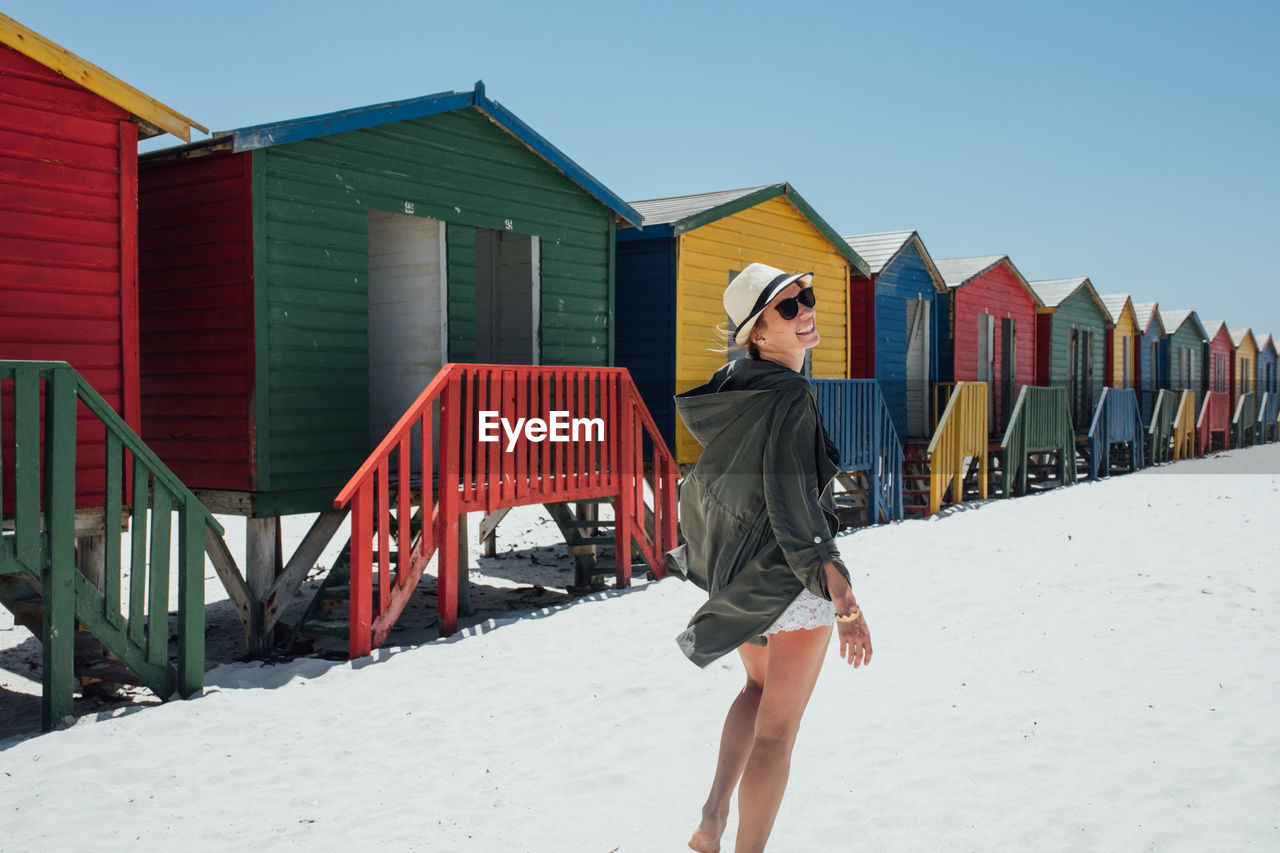 Woman standing at beach against colorful huts and clear blue sky during sunny day