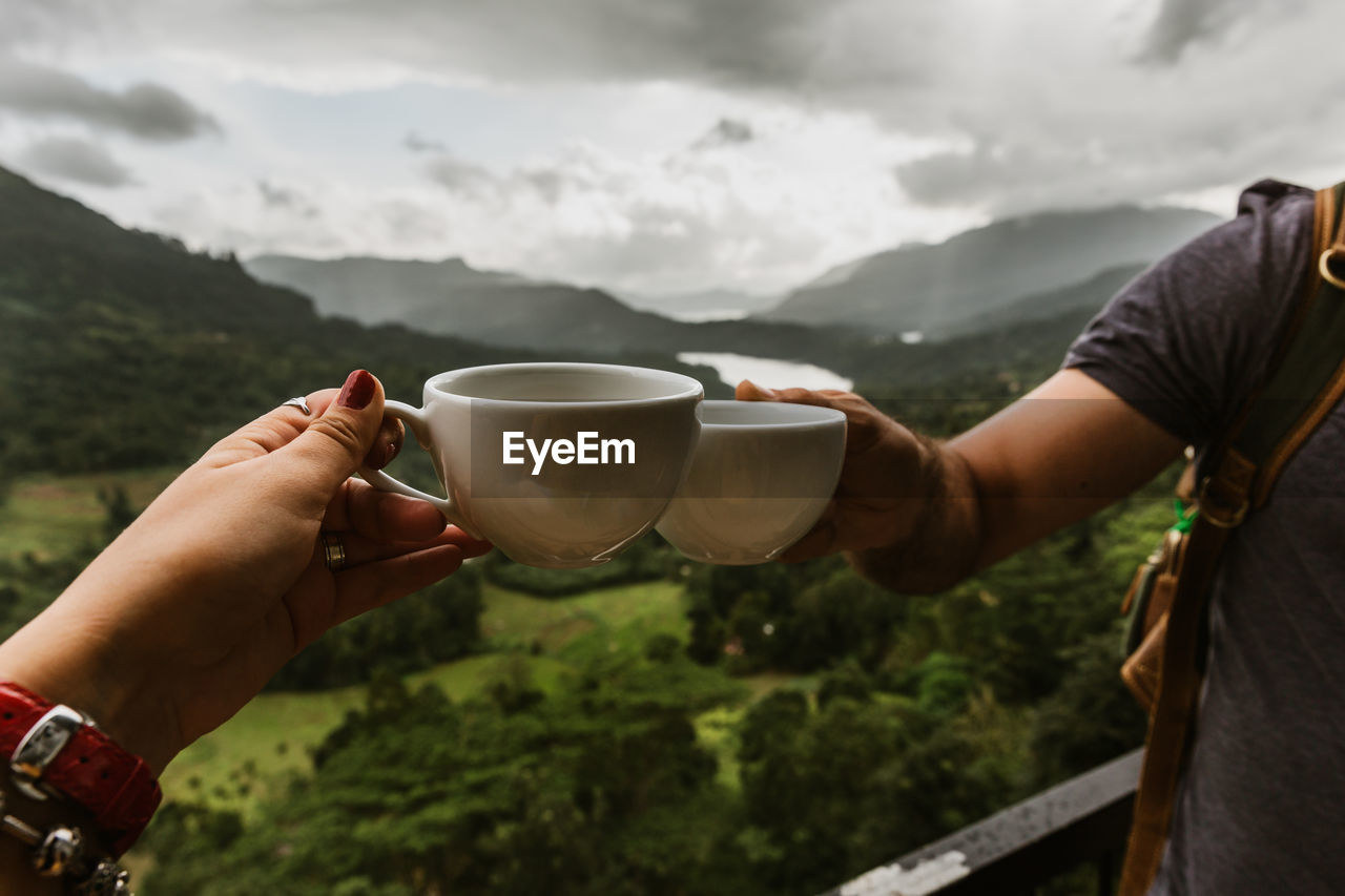 Cropped hands of couple toasting coffee cups against mountain