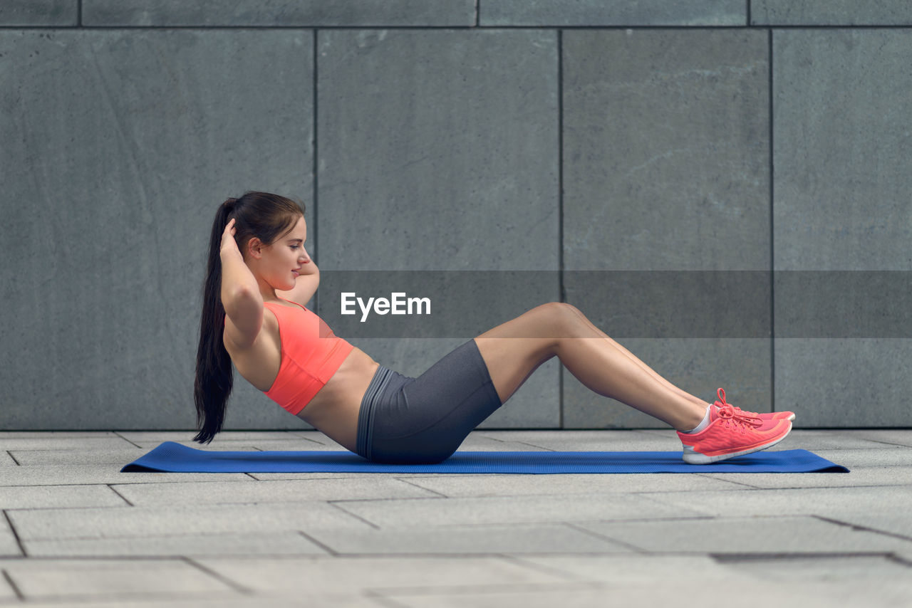 Side view full length of young woman exercising on mat against wall