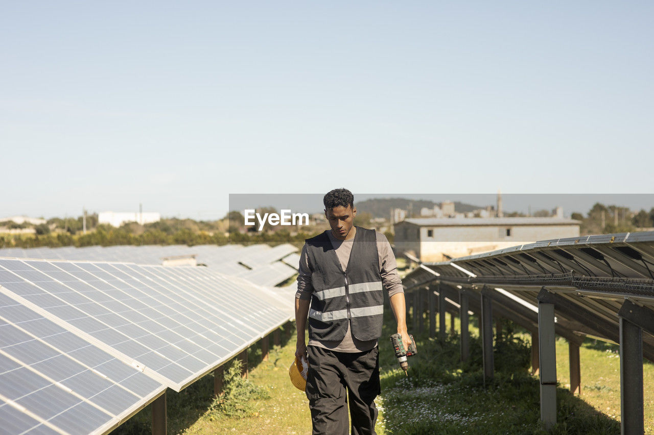 Young male maintenance engineer holding drill while walking amidst solar panels at power station