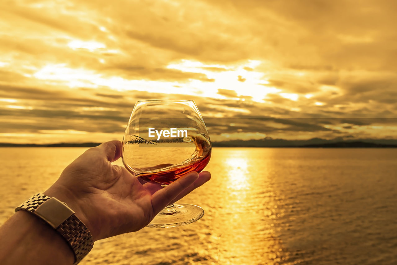 Swirling a alcoholic drink in a crystal snifter glass during golden sunset over the sea.