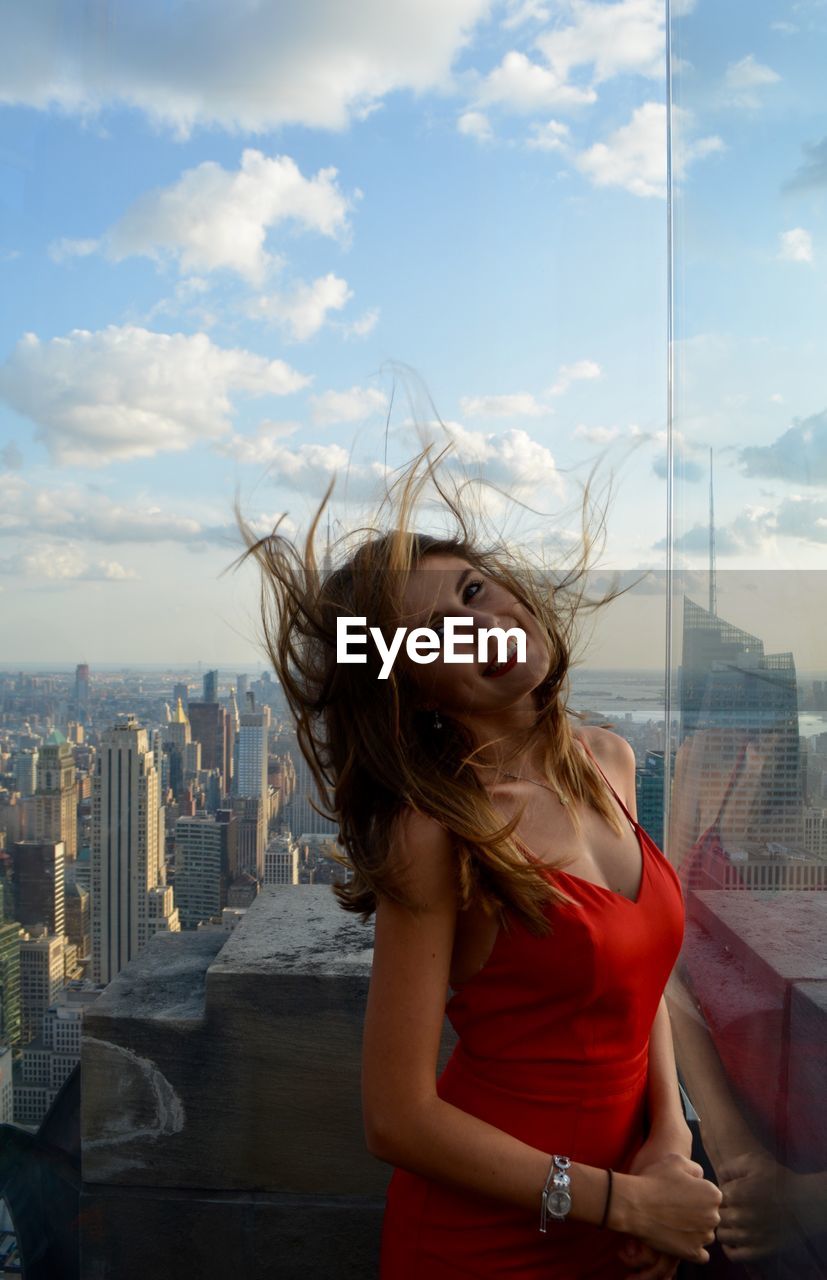 Portrait of cheerful woman with tousled hair standing in city against sky