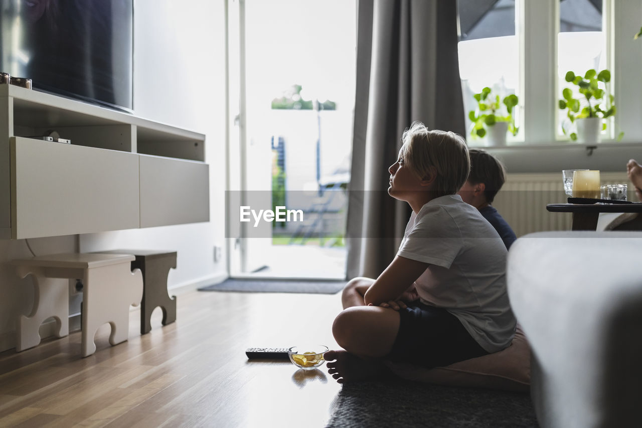 Siblings watching tv while sitting on carpet in living room at home