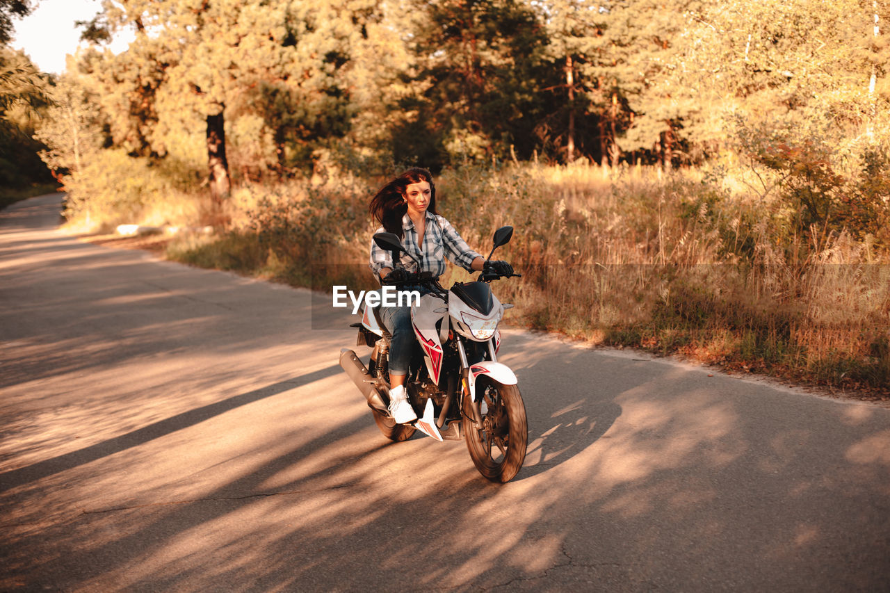 Young confident woman riding motorcycle on country road in forest