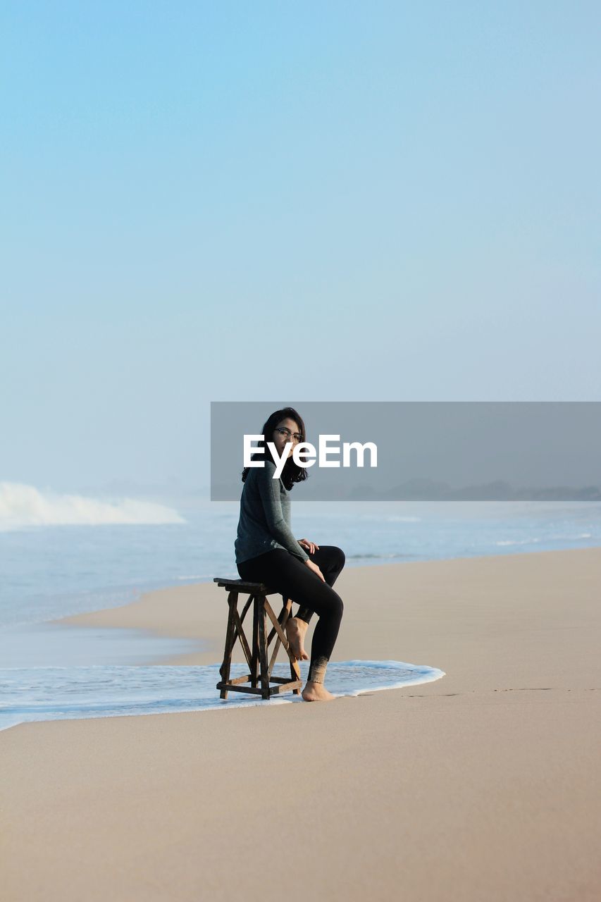 Side view of woman sitting on seat at beach against blue sky