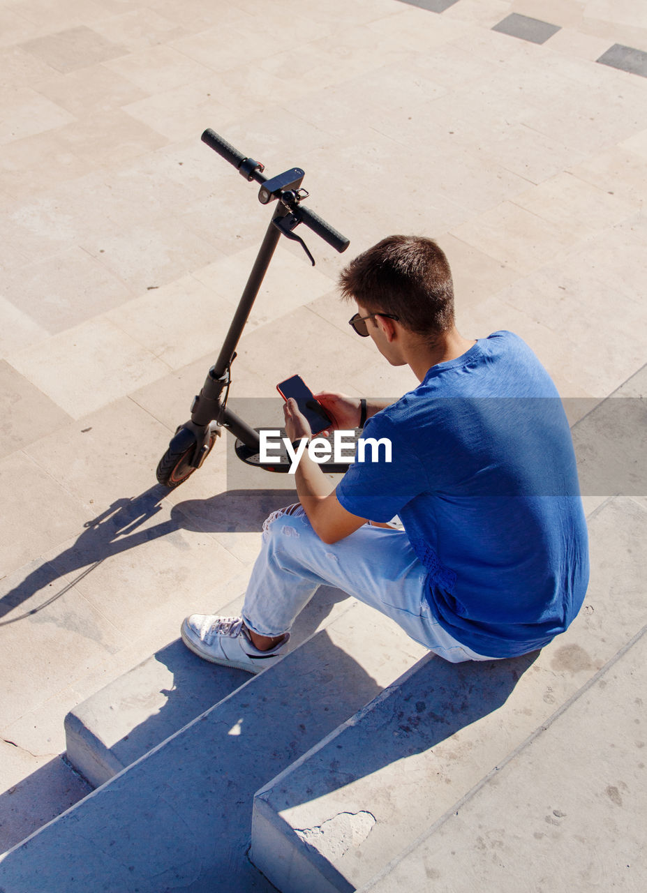 Young caucasian man with smartphone sitting outdoor near electric scooter and using smartphone