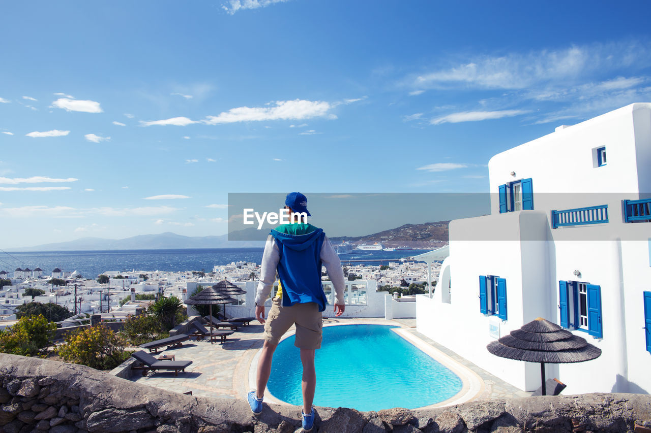 Rear view of man looking at sea while standing on retaining wall by house