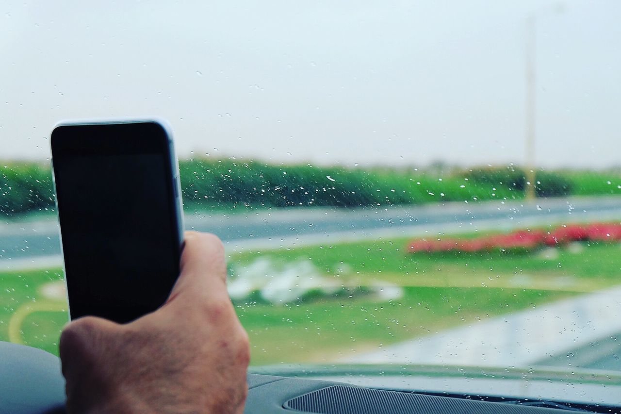 Cropped hand of man holding smart phone against wet windshield in car