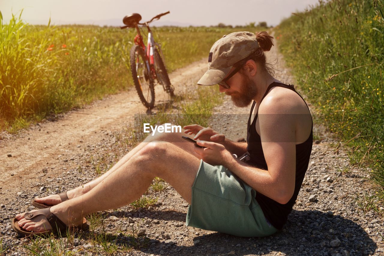 Side view of man using mobile phone while sitting on land by bicycle
