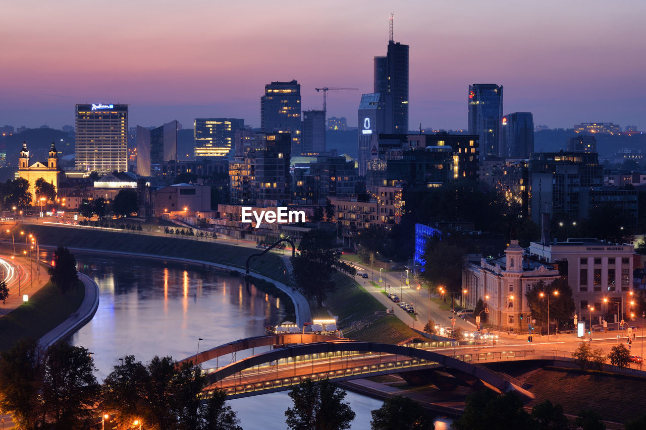 Illuminated bridge over river by buildings against sky at night