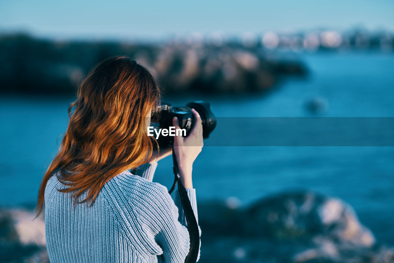 REAR VIEW OF WOMAN PHOTOGRAPHING WITH SEA IN BACKGROUND