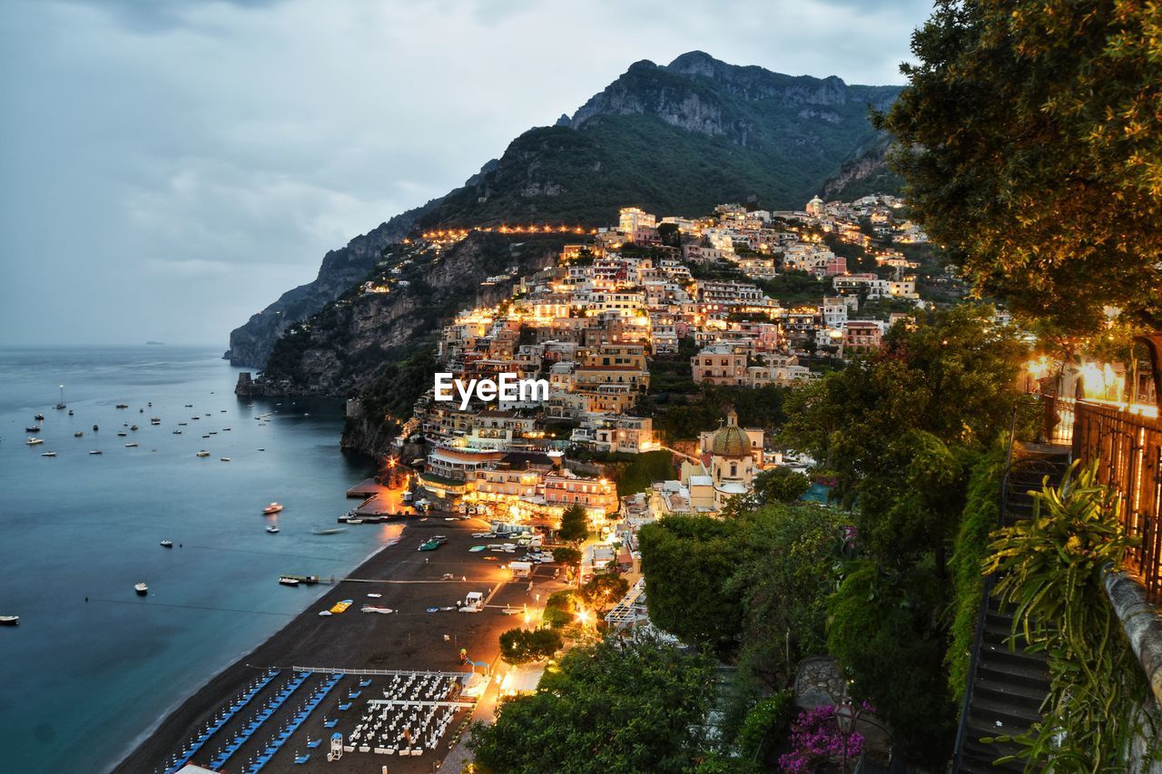 HIGH ANGLE VIEW OF ILLUMINATED TOWNSCAPE BY SEA AGAINST SKY