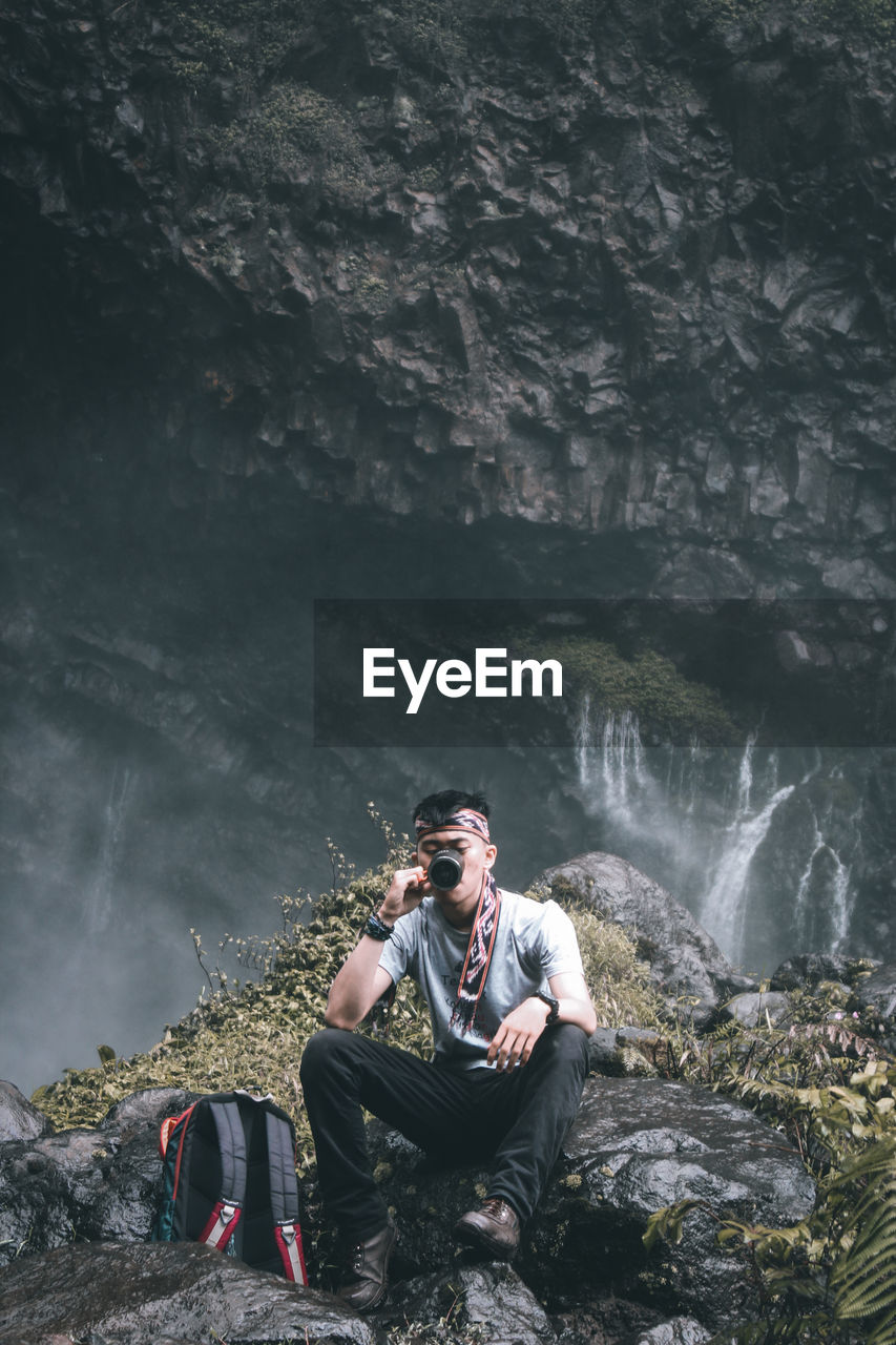 Young man sitting on rock against waterfall