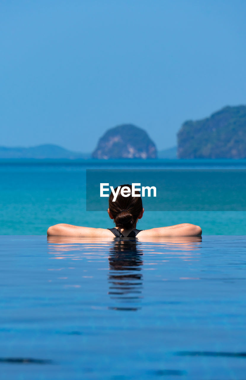  young woman wearing bikini standing in blue infinity swimming pool looking at the view of ocean 