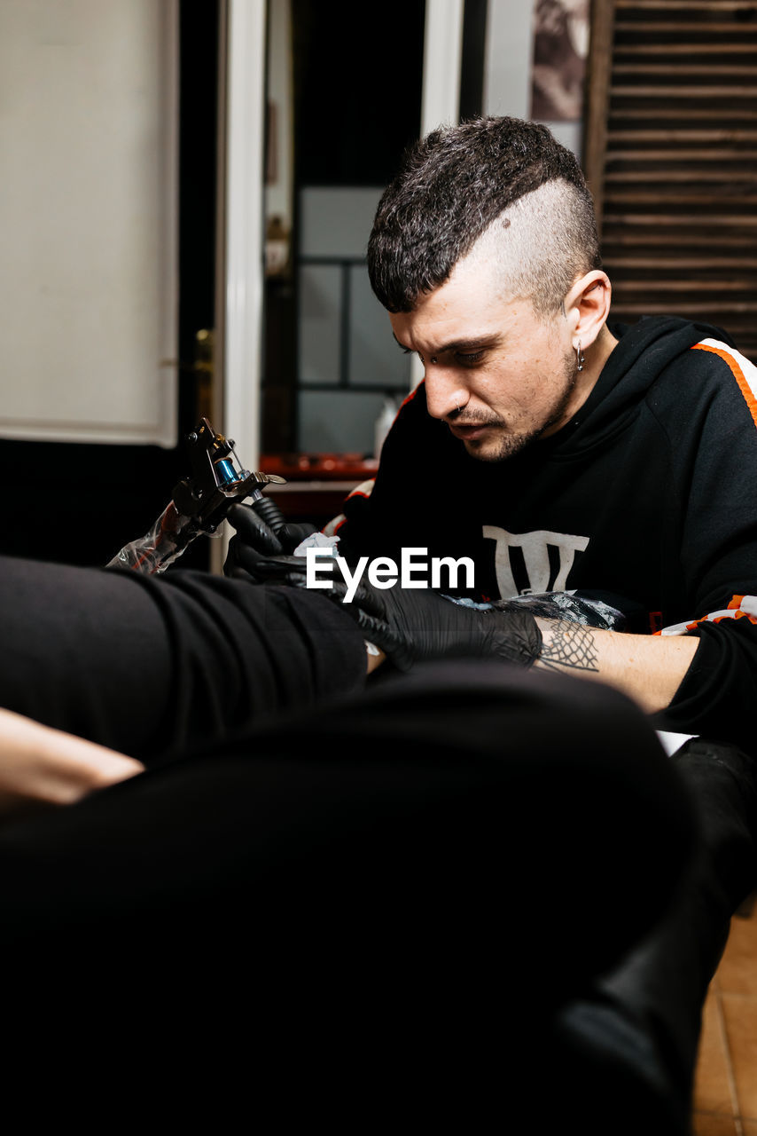 Stylish man with piercing using tattoo machine to make tattoo on leg of crop customer during work in salon