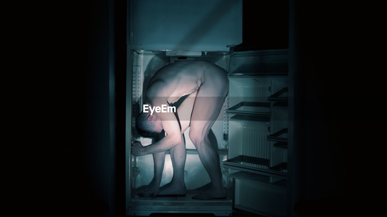 Full length of naked man standing in refrigerator in darkroom