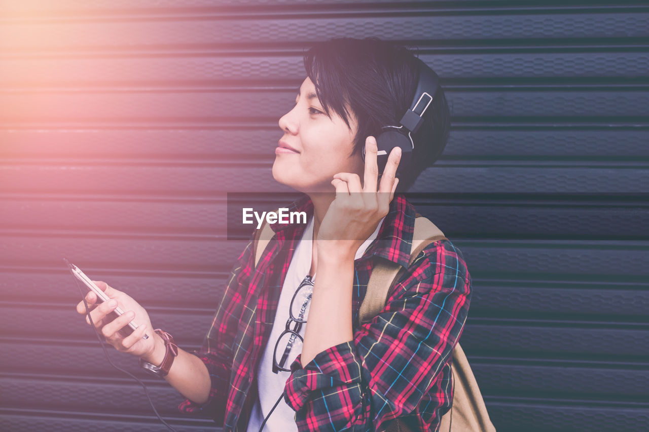 Smiling young woman listening music on headphones while standing against shutter