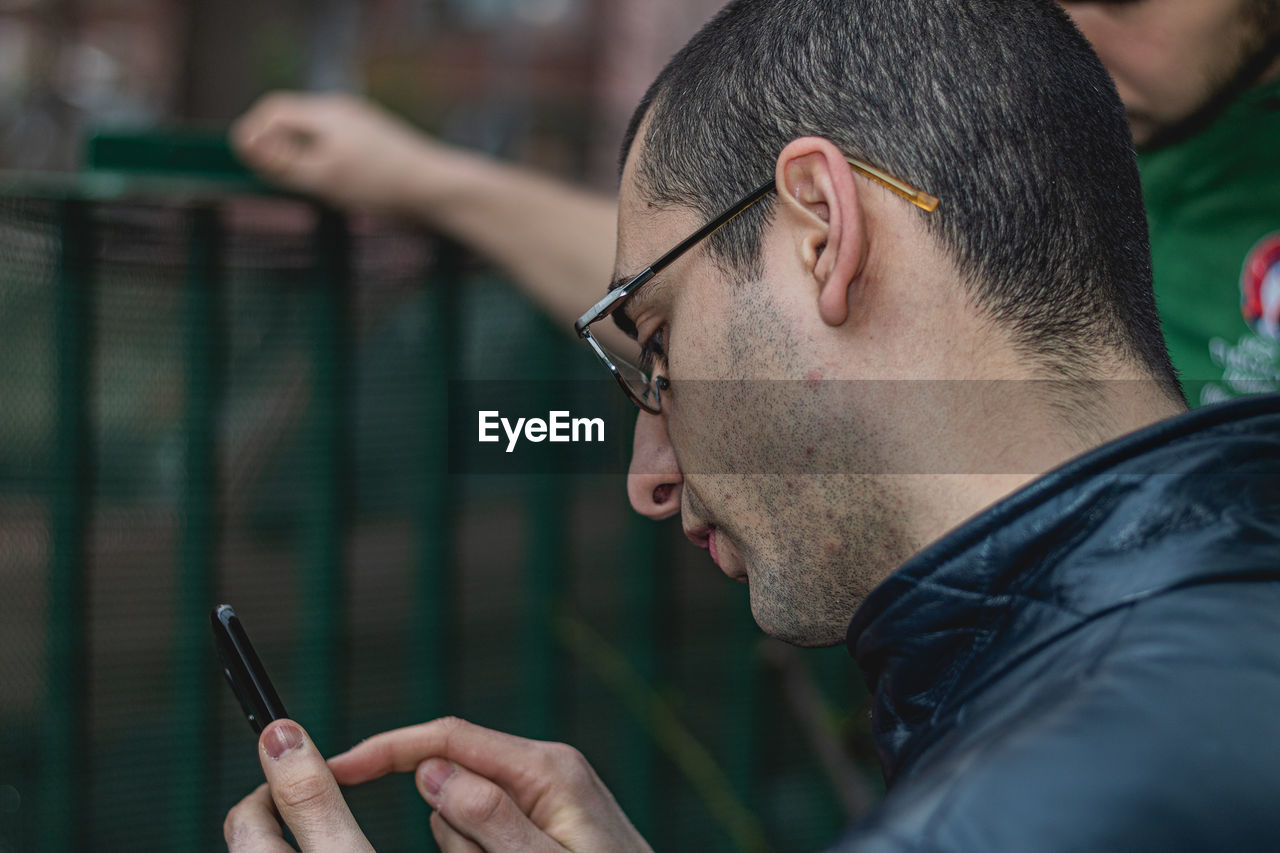 CLOSE-UP PORTRAIT OF YOUNG MAN USING SMART PHONE OUTDOORS