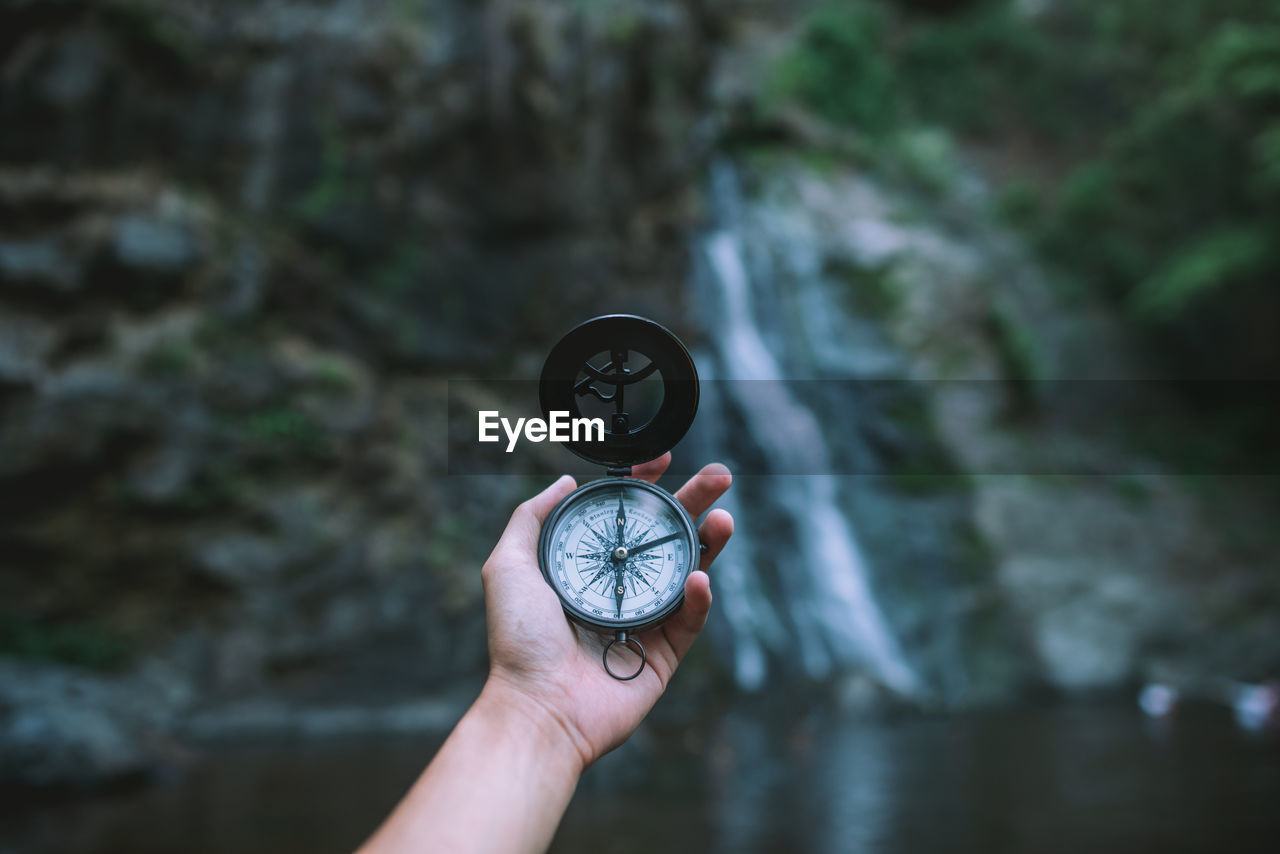Cropped hand of person holding navigational compass against waterfall