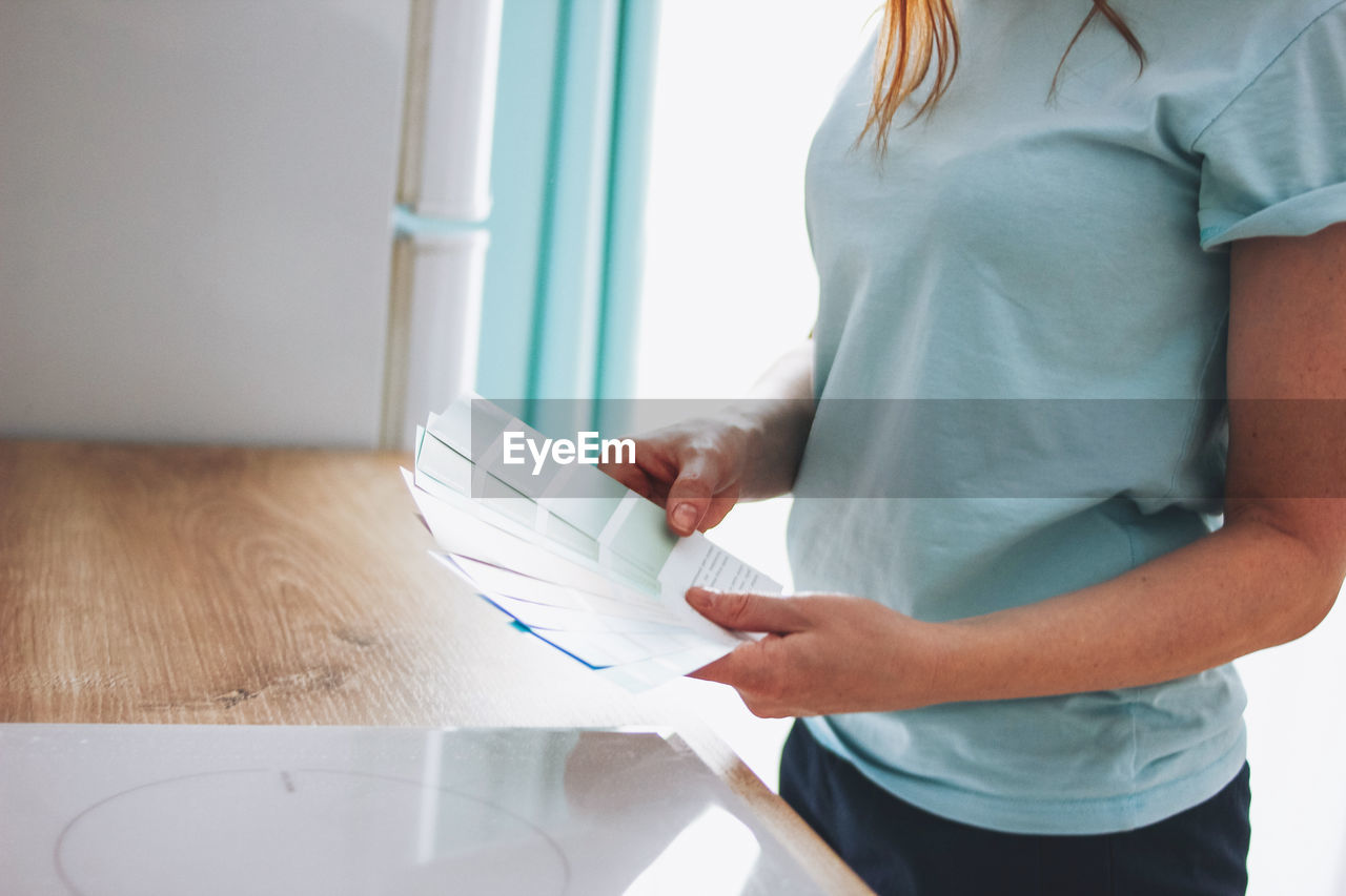 MIDSECTION OF WOMAN HOLDING BOOK WHILE STANDING AT TABLE